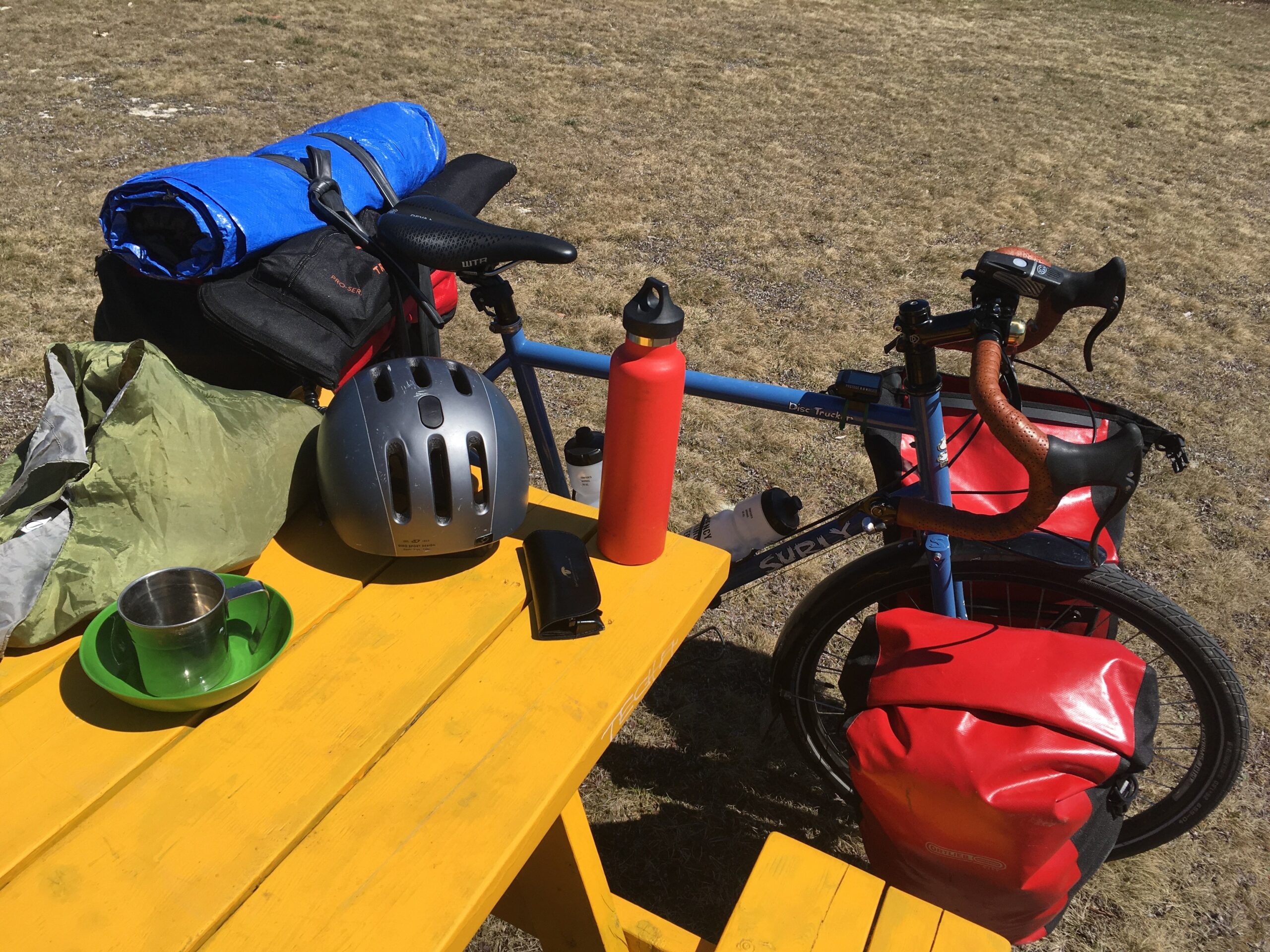 A blue bike with red panniers next to a yellow picnic table.