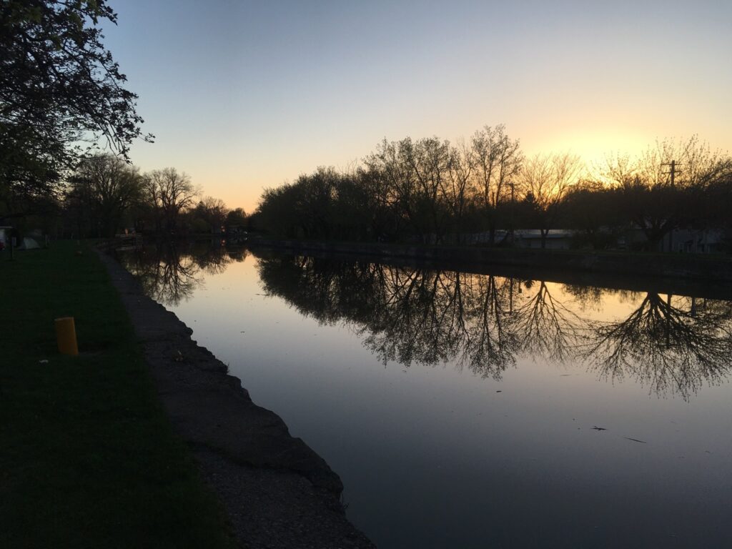 A an evening view of the Erie Canal. The water is smooth as glass, and the trees, still bare late in the spring, reflect off of it.