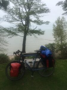 A touring bike loaded with gear, leaning on a park bench with Lake Erie in the background on a cloudy misty day.