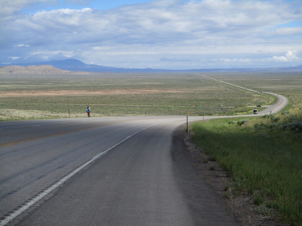An eastbound Trans America cyclist approaches up this hill in Wyoming, north of Rawlins. There is a wide open valley at the bottom of the hill with a road crossing it. Mountains are in the distance.