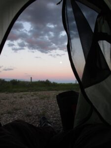 A view out a one-person backpacking tent. A wire fence and sage brush can be seen pass a gravel drive. The moon is in the sky.
