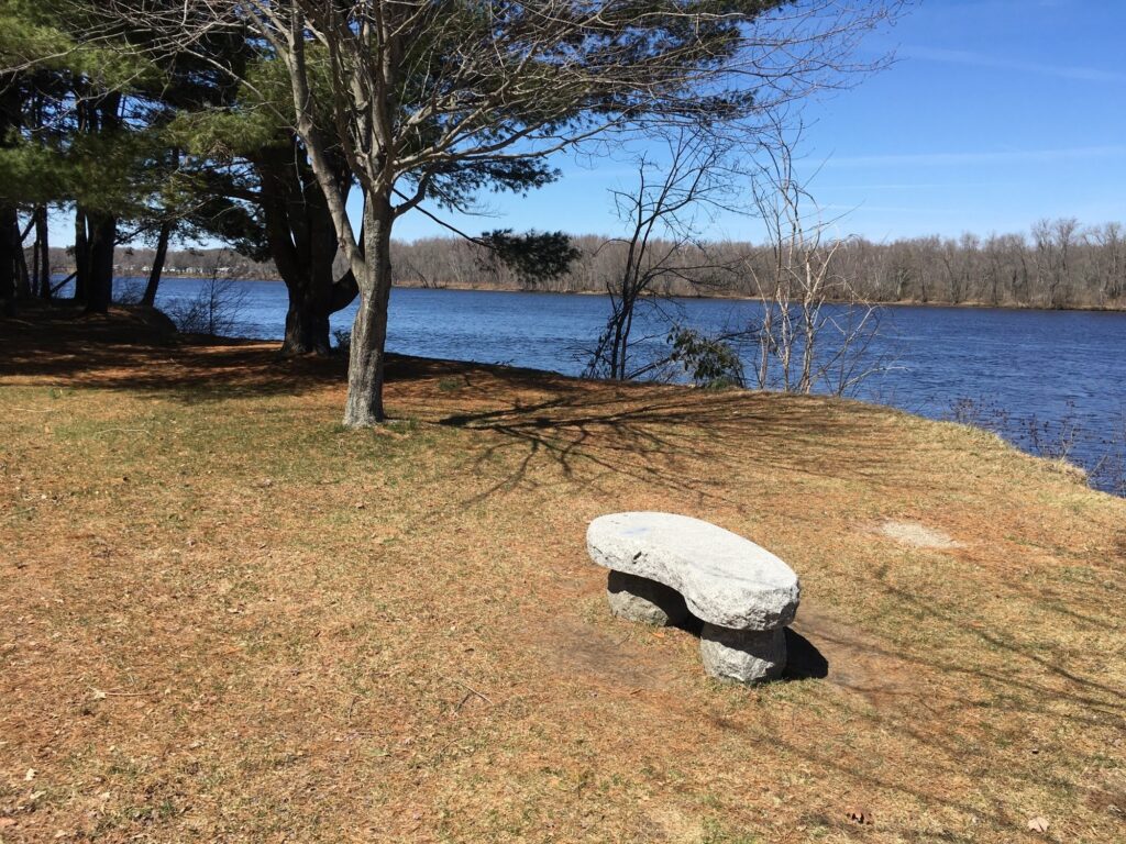 A small stone bench along Brunswick bike pathe with the Androscoggins River and a blue sky in the background.